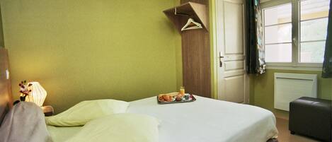 Una cama bien hecha con una bandeja de desayuno, un abrigo colgado y una lámpara de noche en una habitación con paredes verdes y una ventana.