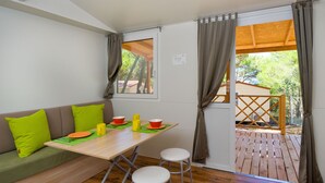 Una habitación con una mesa de madera, cojines verdes y vistas a una terraza de madera y árboles.