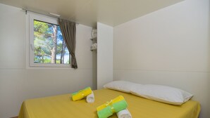 Una cama individual con una colcha amarilla, dos toallas enrolladas, una almohada blanca y una ventana con vistas a árboles.