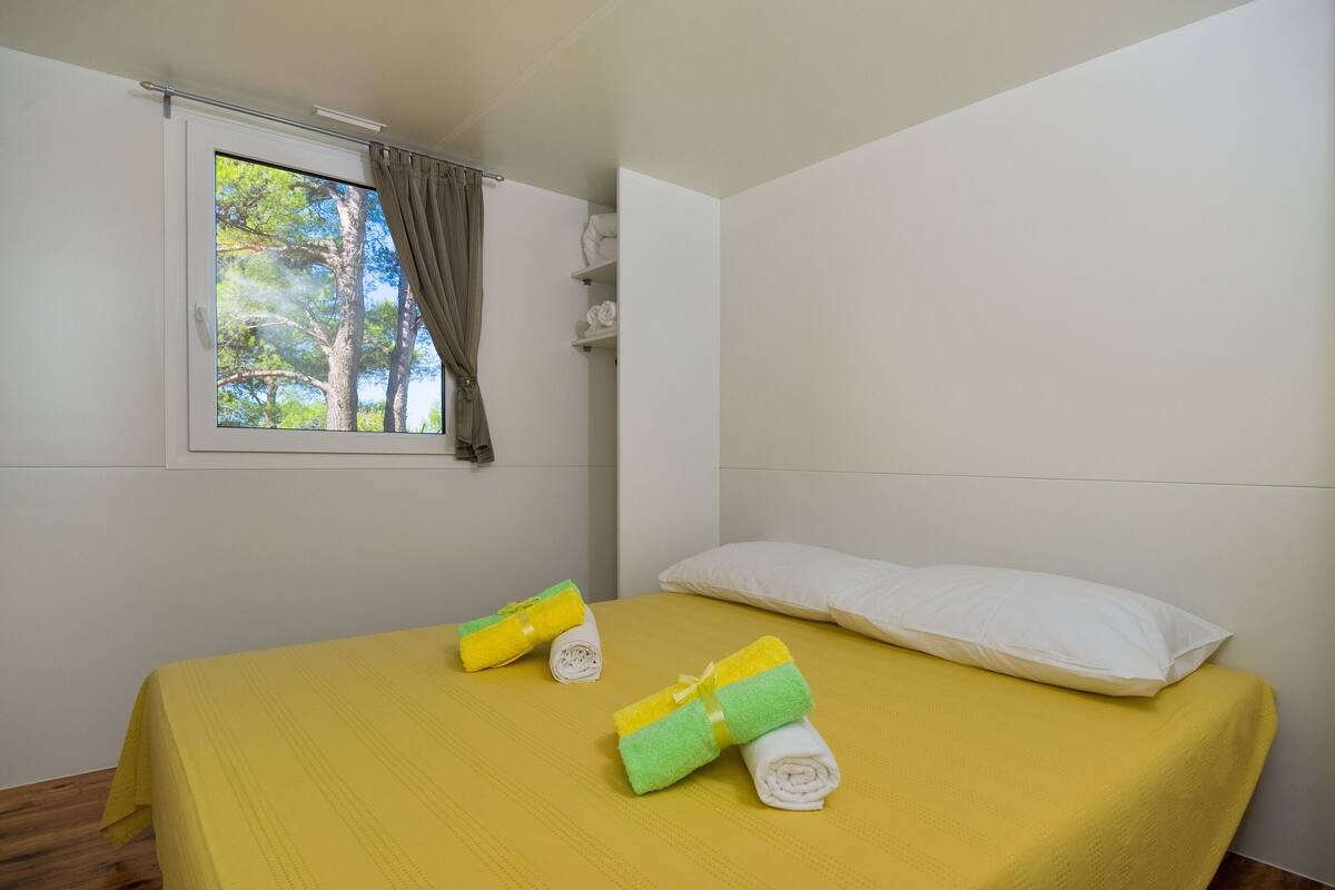 Un lit simple avec une couvre-lit jaune, deux serviettes enroulées, un oreiller blanc et une fenêtre donnant sur des arbres.
