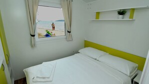 Un dormitorio con cama, una ventana con cortinas, una estantería con una planta y vistas a la playa.