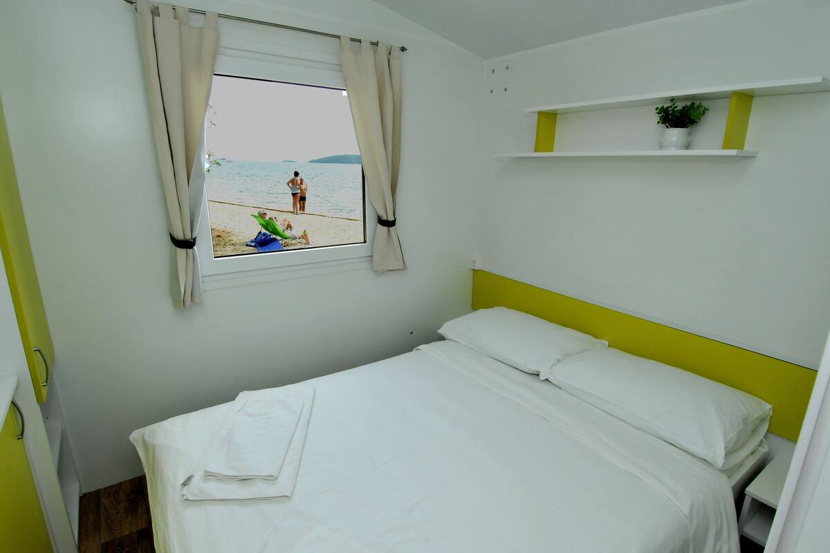 A bedroom with a bed, a window with curtains, a shelf with a plant, and a view of the beach.