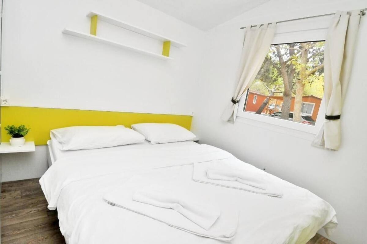 A neatly made bed with white bedding, a yellow headboard, and a window with curtains.