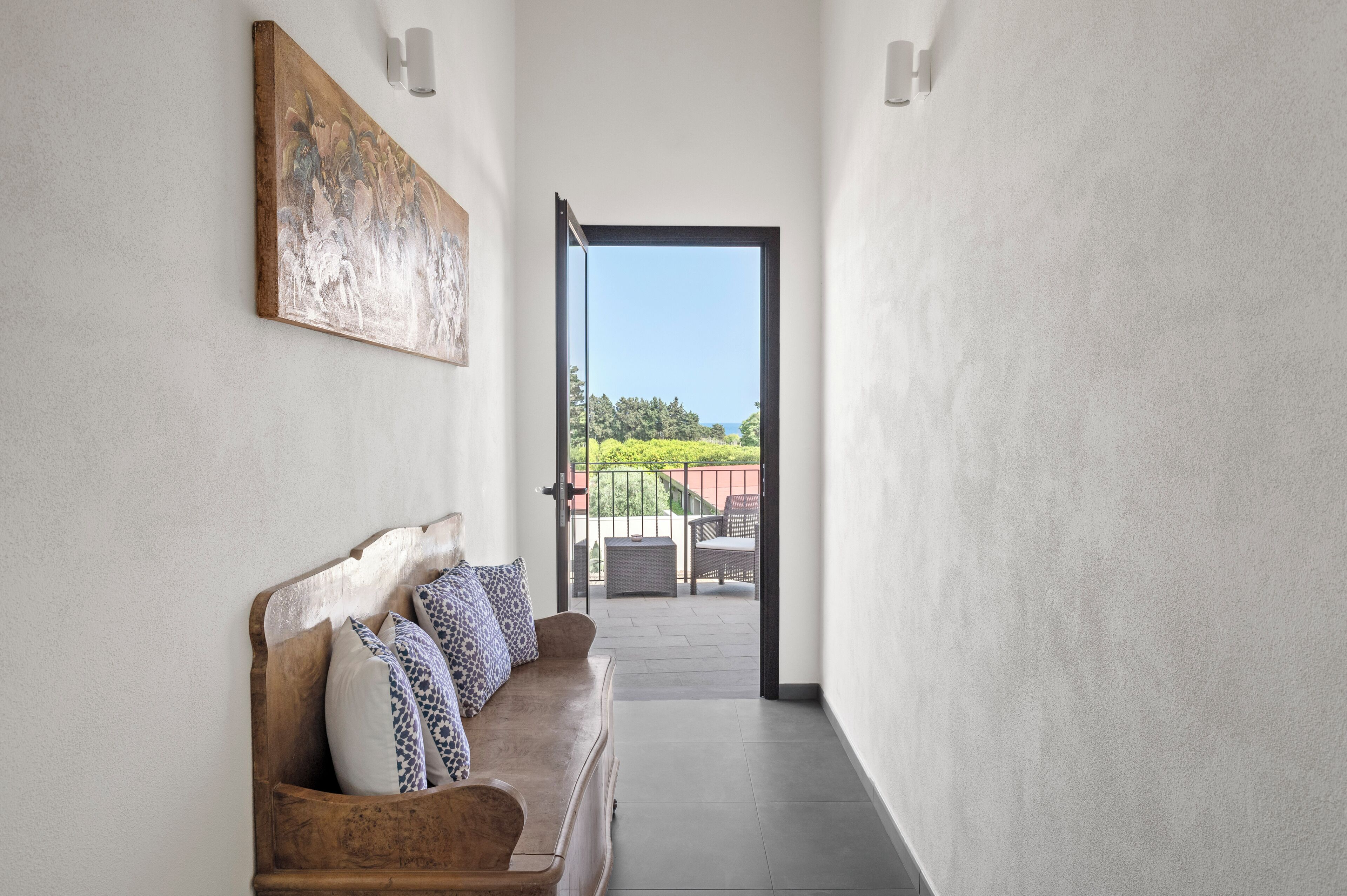 Un couloir étroit, avec un banc en bois et des coussins à motifs, qui mène à un balcon aménagé avec du mobilier d’extérieur.