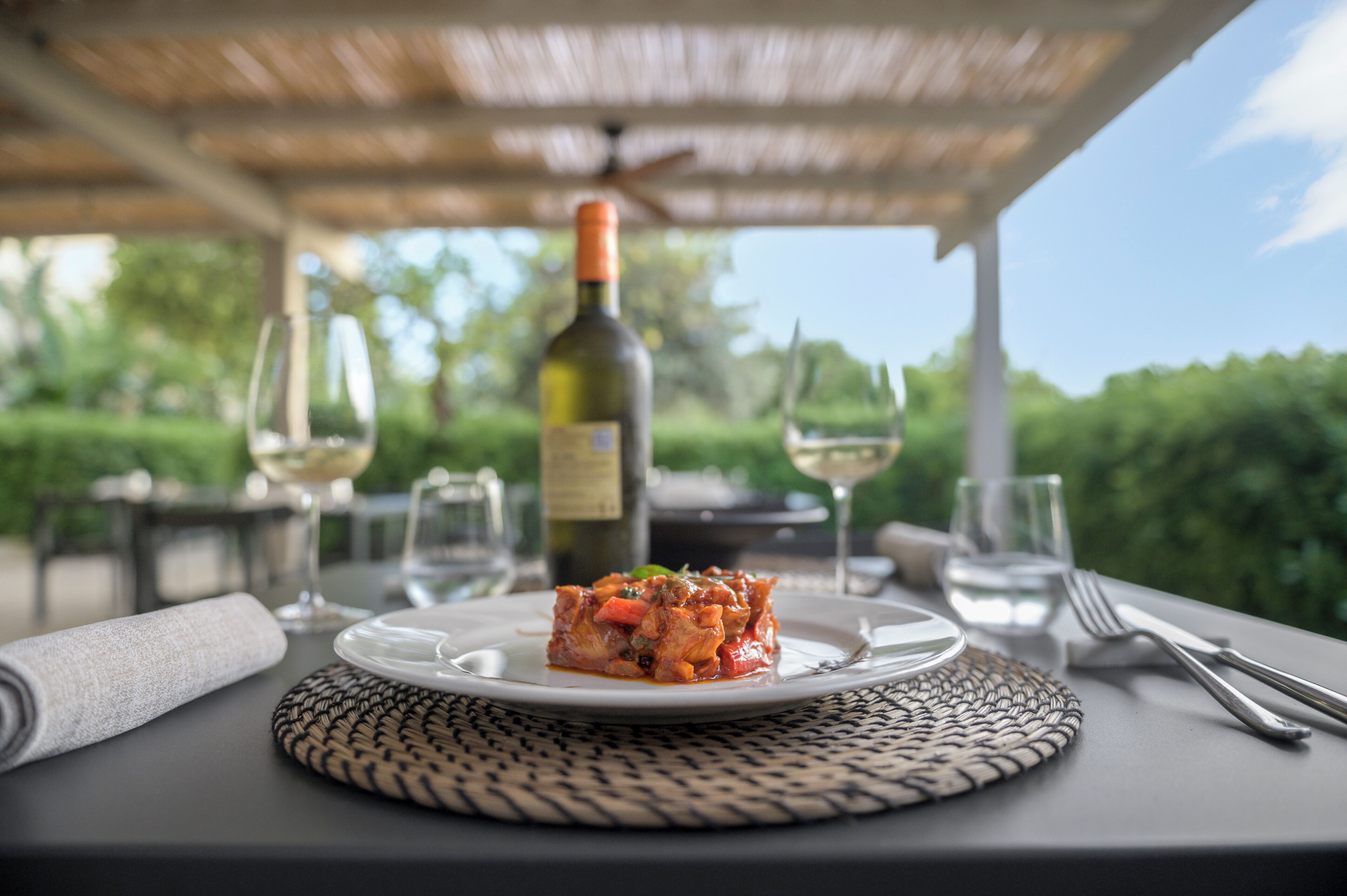 A table set outdoors with a plate of food, a bottle of wine, and glasses.