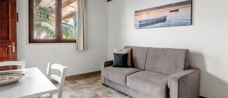 A room with a beige sofa, a white table with a bowl, a window with curtains, and a painting of a boat on water.