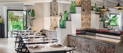 A restaurant with multiple tables, hanging pendant lights, and a wine bar with bottles.