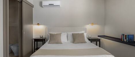 Ein Hotelzimmer mit Bett, Nachttischen, einem Regal mit Büchern, einer Klimaanlage und einer Tür zu einem Badezimmer.