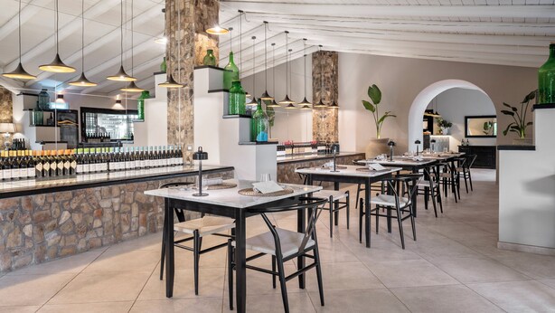 Un restaurante con varias mesas y sillas, una zona de bar con botellas, y lámparas colgantes.