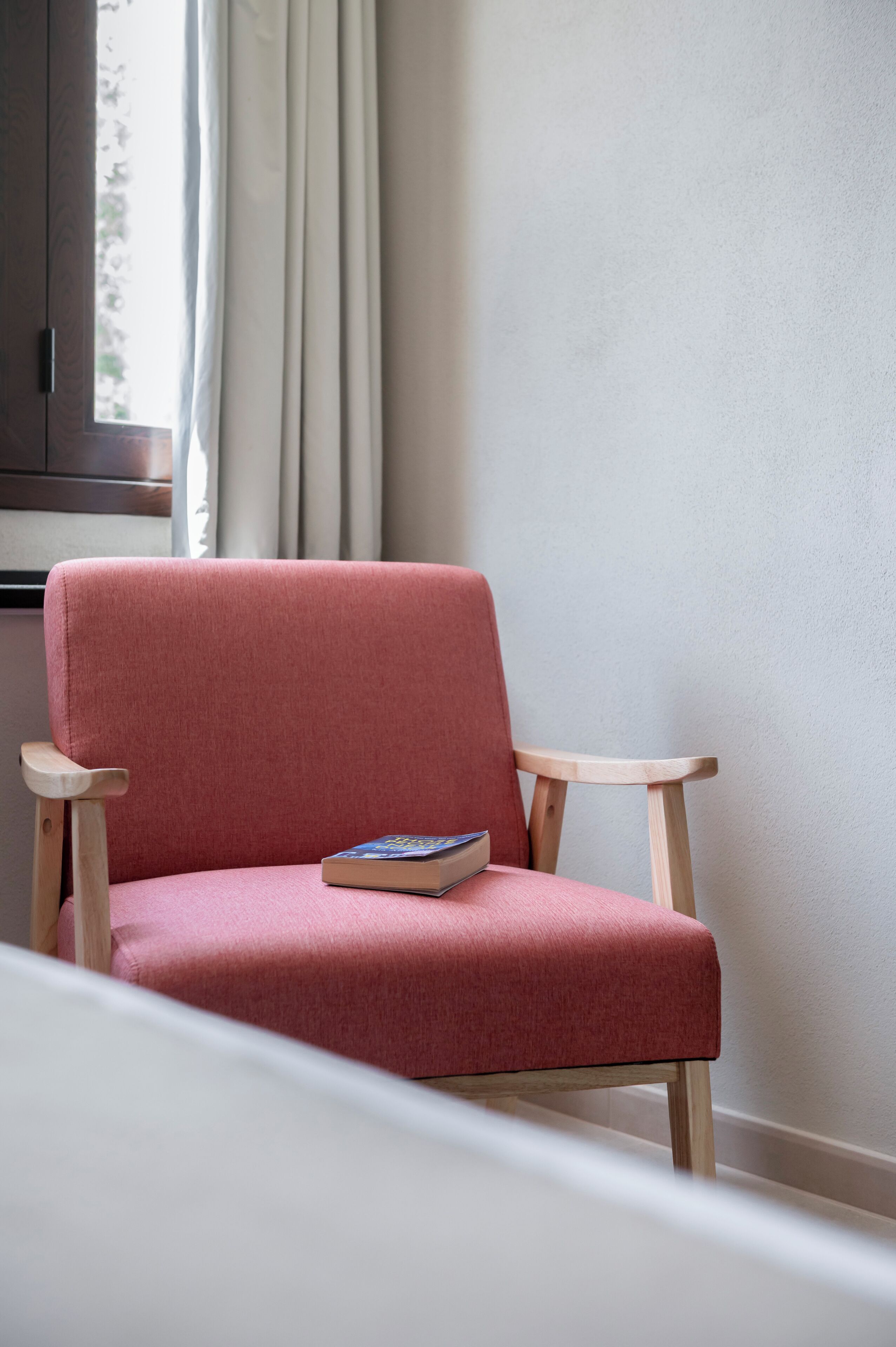 A red upholstered chair with a book on it, positioned near a window with curtains.