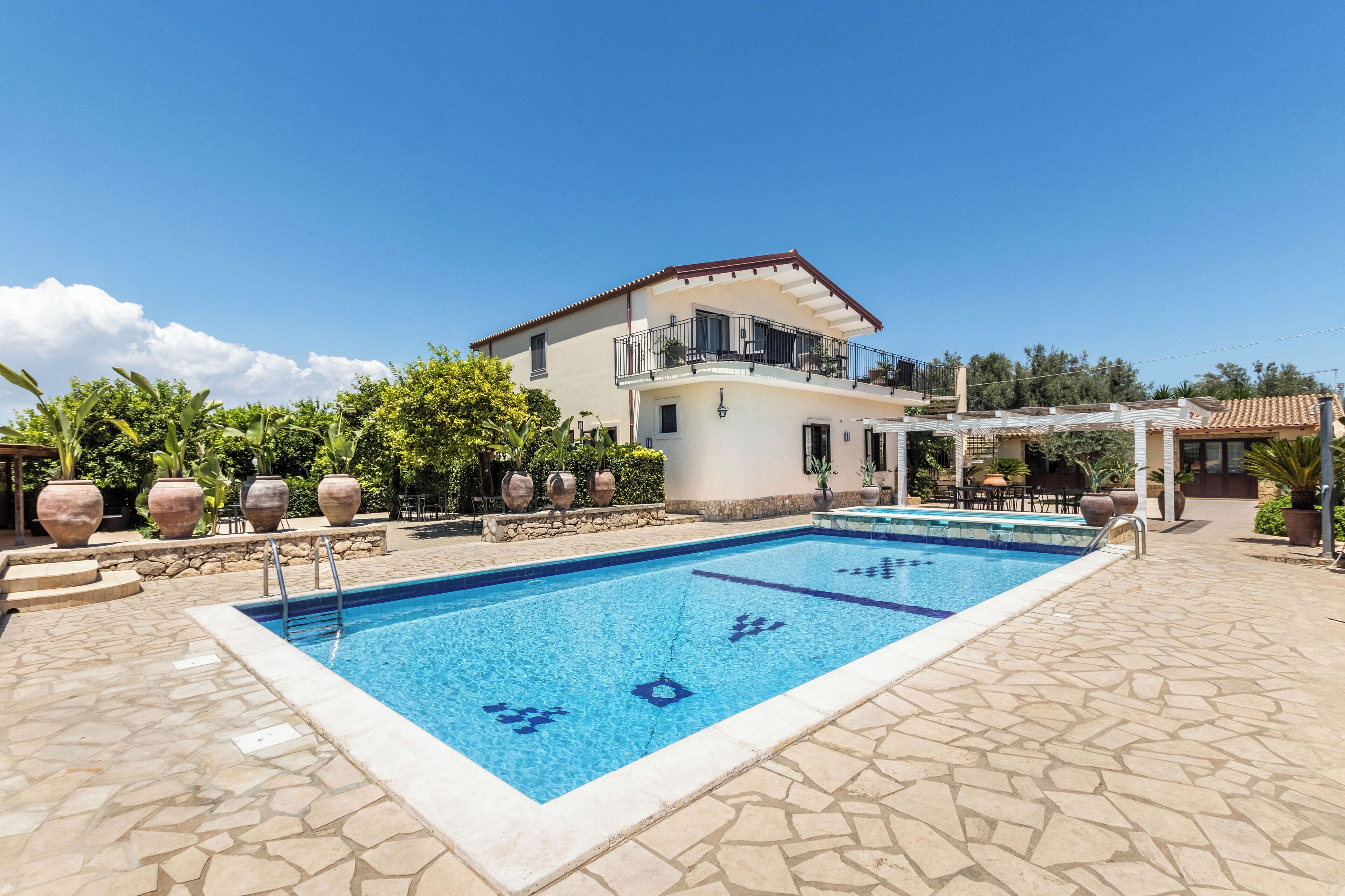 A swimming pool with a tiled deck, a two-story building with a balcony, and potted plants around the pool area.