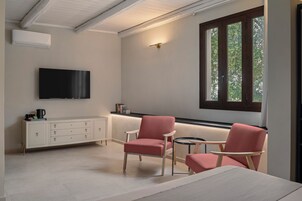 Uma sala de estar moderna com uma televisão de tela plana, um armário branco, duas cadeiras cor-de-rosa, uma mesa redonda pequena e uma janela grande com vista para árvores.