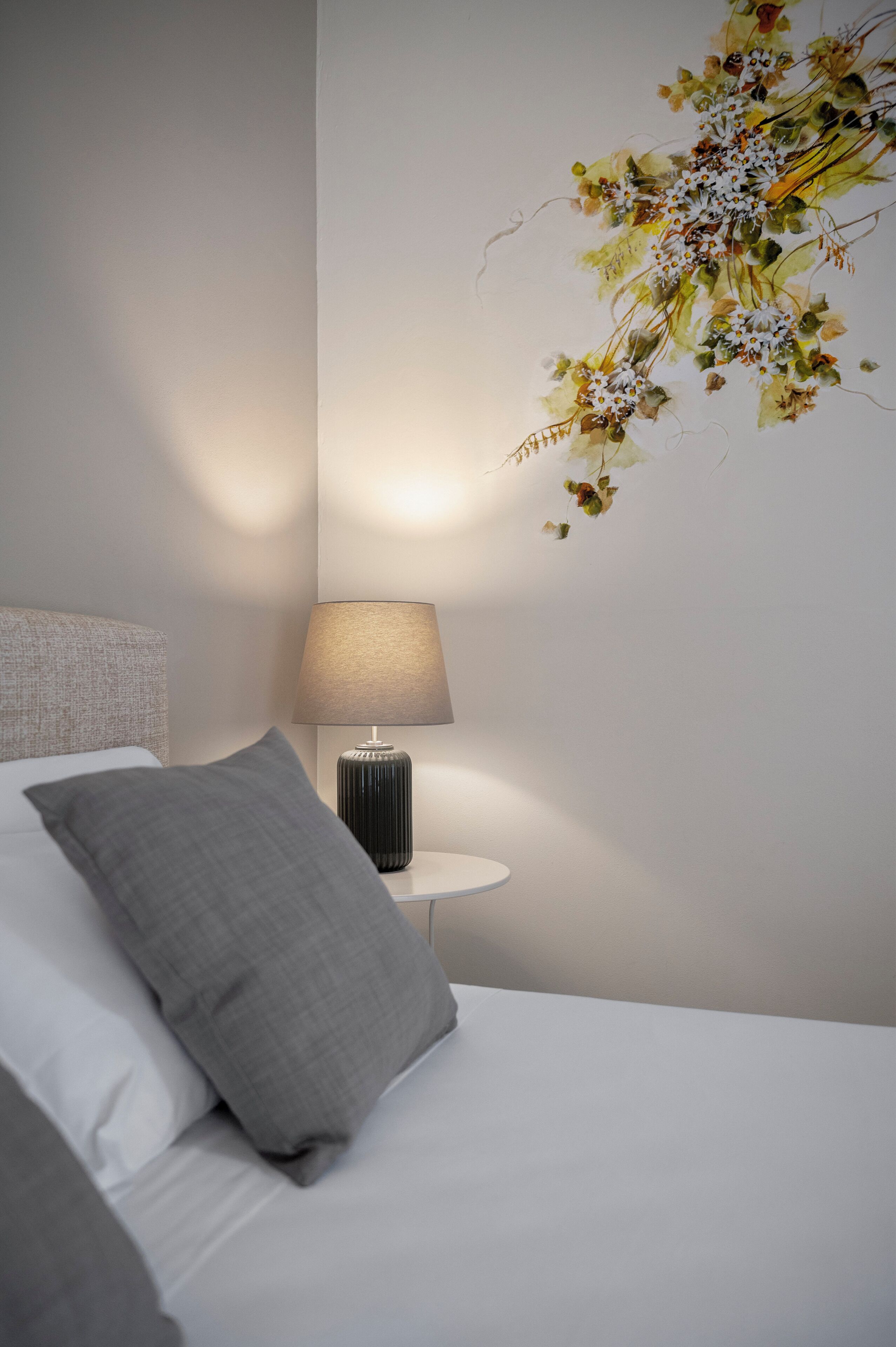 A neatly made bed with a grey pillow, a bedside lamp, and a wall-mounted floral decoration.