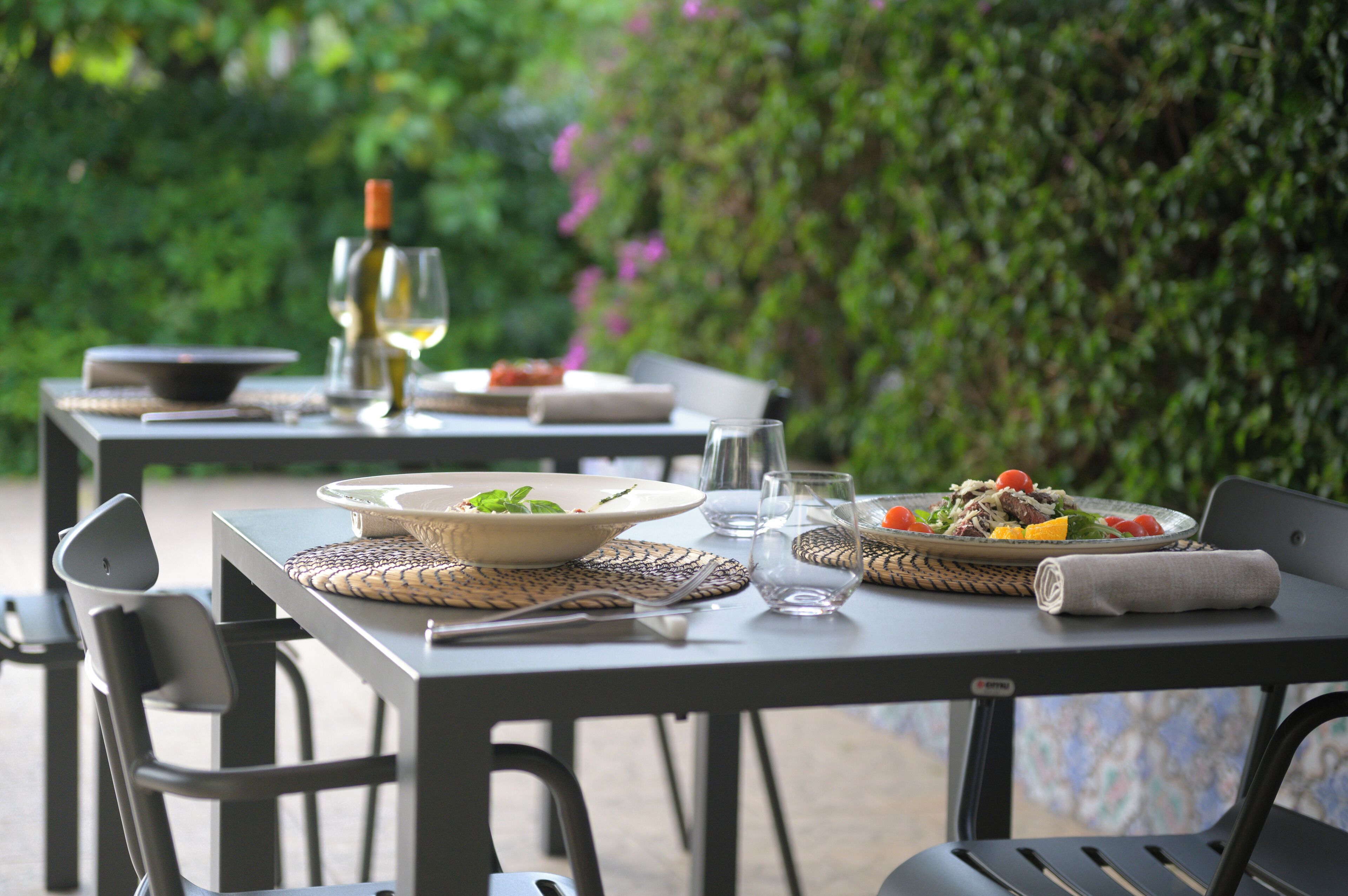 Area esterna con zona pranzo, tavoli apparecchiati per due, con tovagliette intrecciate e diversi piatti.