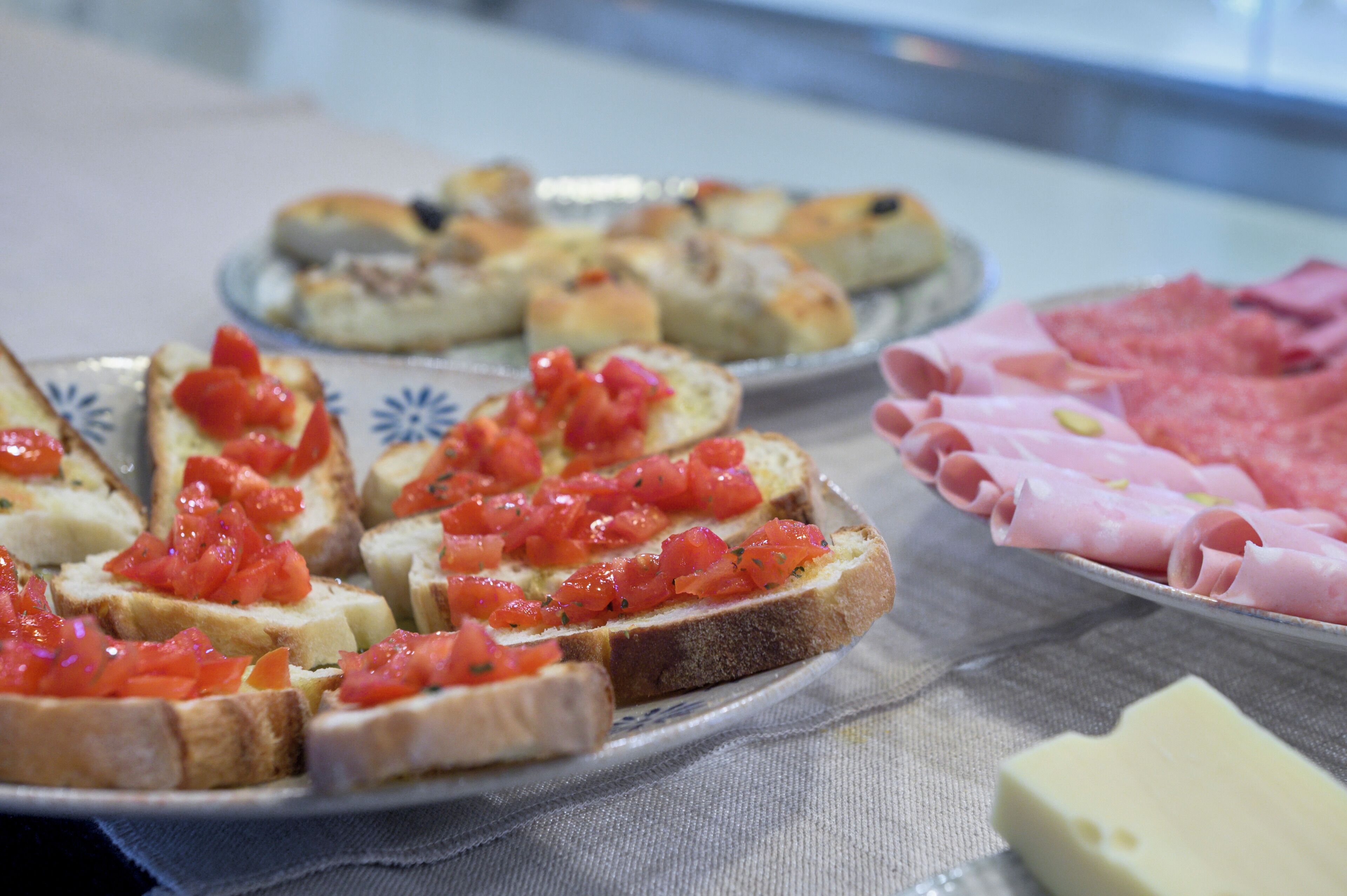 A variety of food items including bread with tomato topping, meat slices, and a block of cheese on a table.