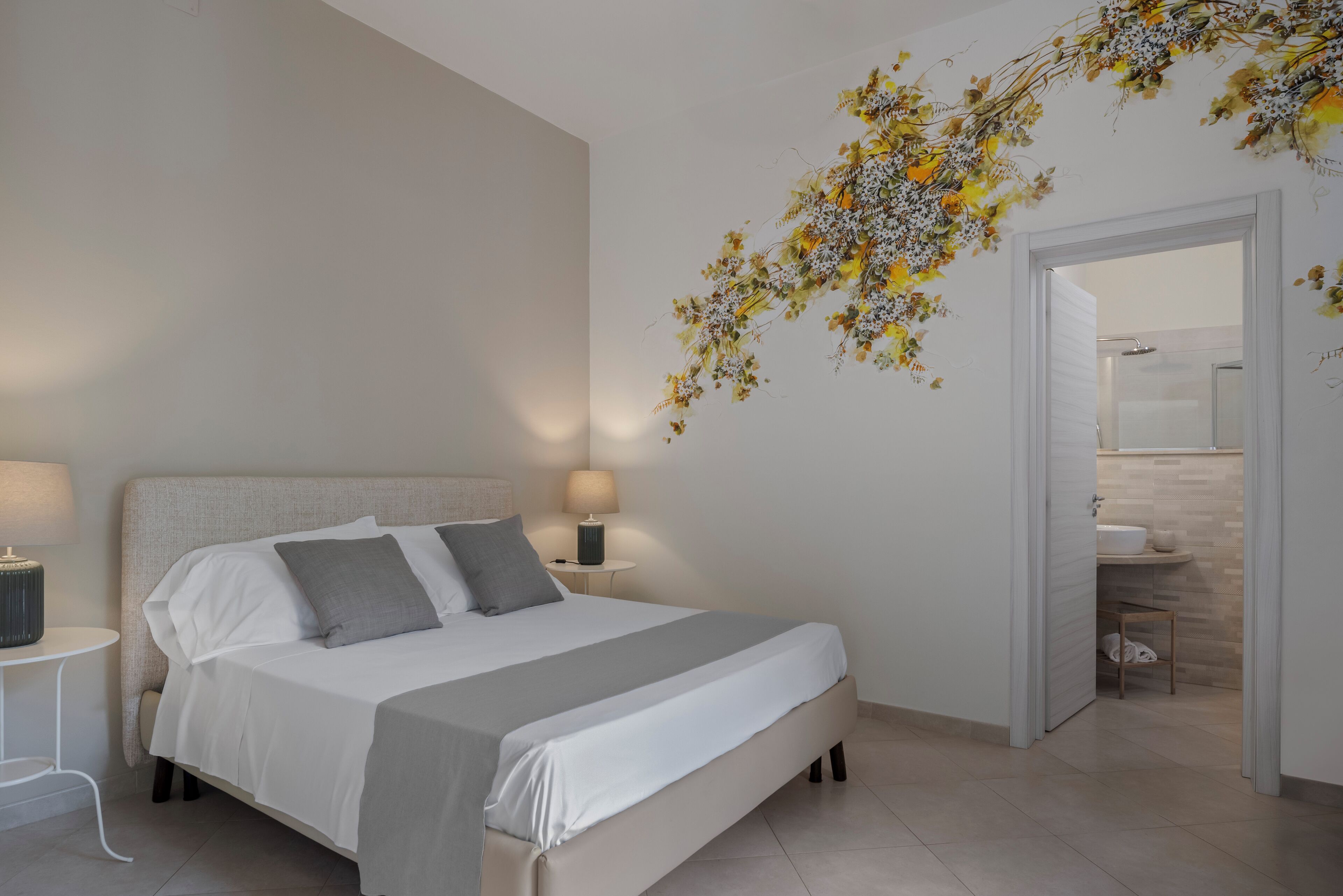 A modern bedroom with a large bed, two bedside tables with lamps, a decorative floral wall piece, and a view into the bathroom.