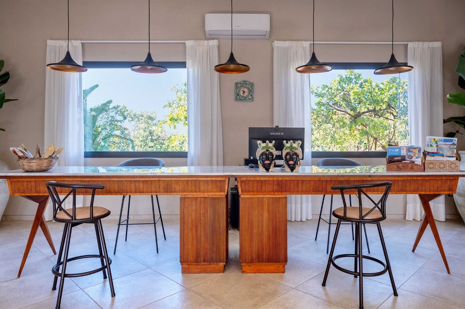 A kitchen with a wooden island, two bar stools, pendant lights, and a view of trees through large windows.