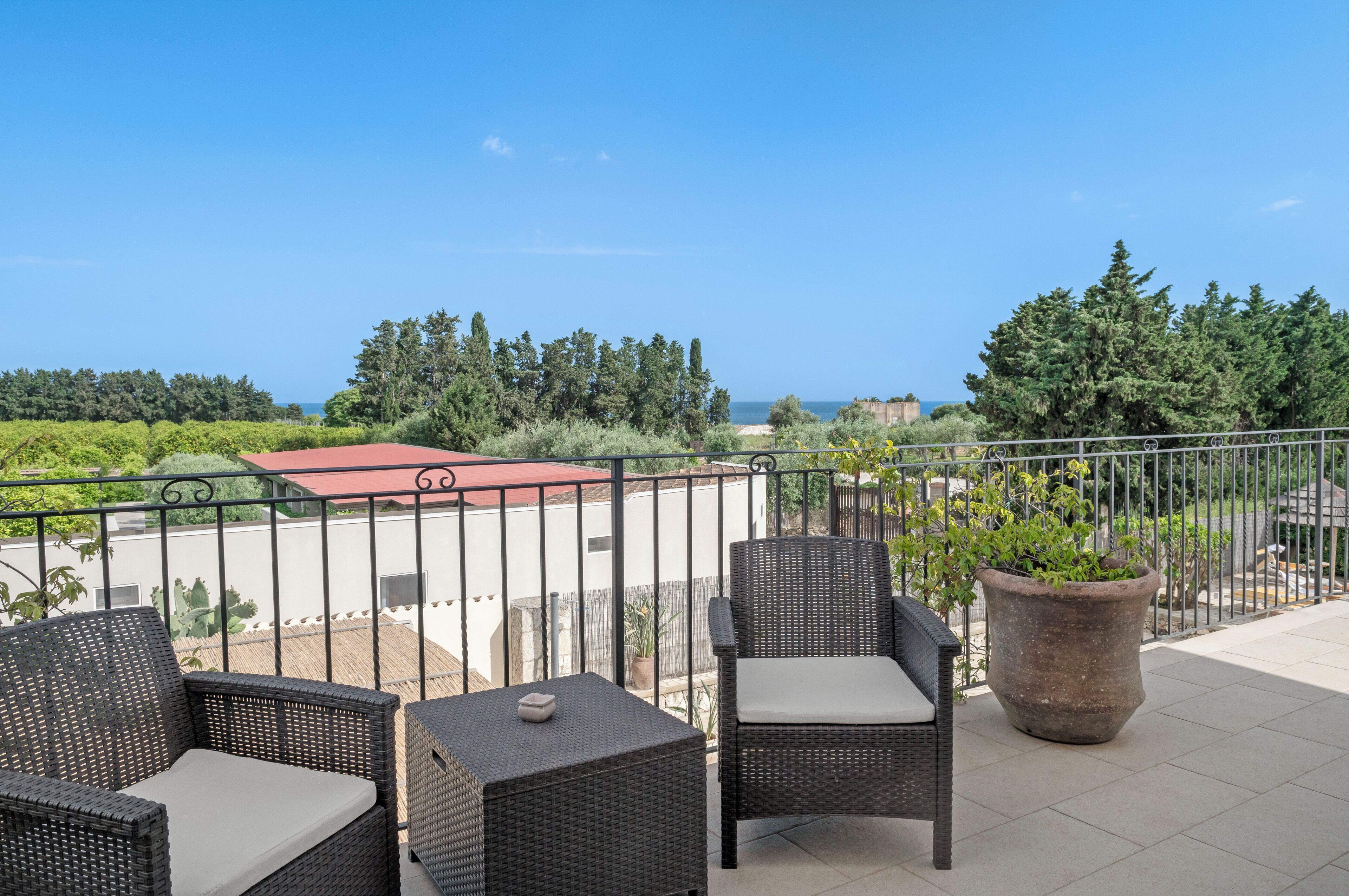 A balcony with wicker furniture, a potted plant, and a view of the sea and greenery.