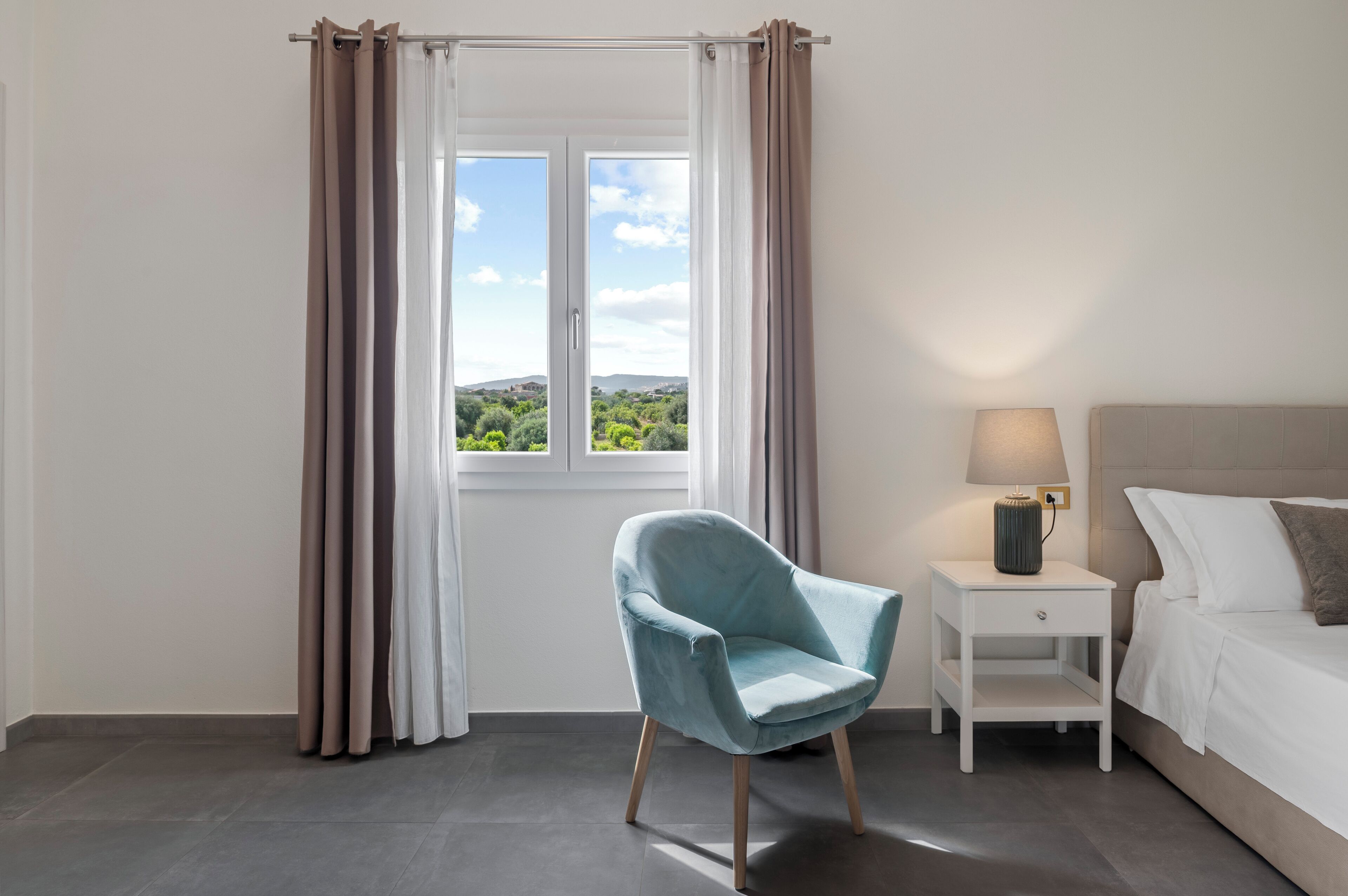 Ein Schlafzimmer mit Bett, Nachttisch, Lampe, Sessel und einem Fenster mit Vorhängen.