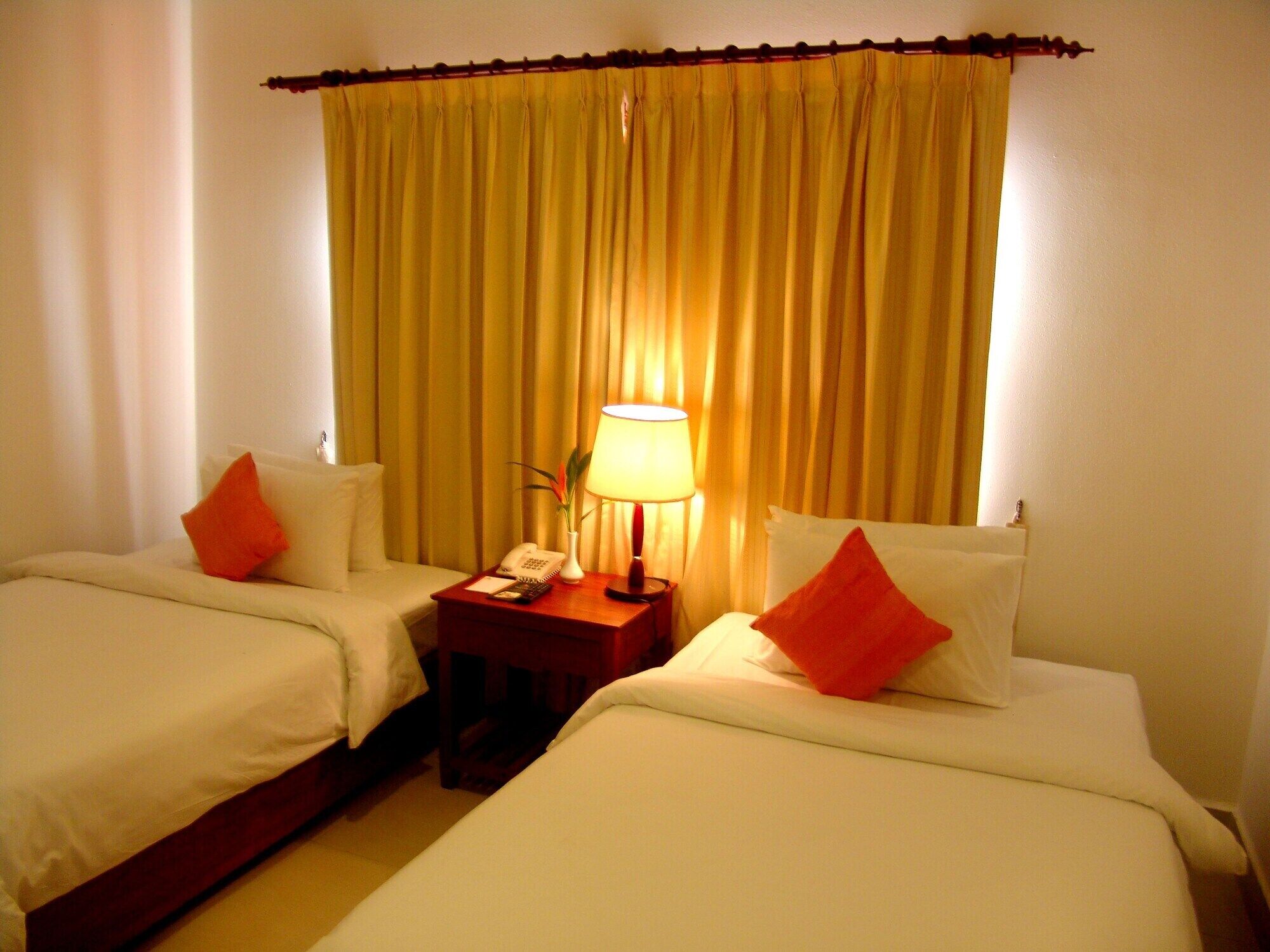 A hotel room with two beds, a nightstand with a lamp, a phone, and a vase with a plant.