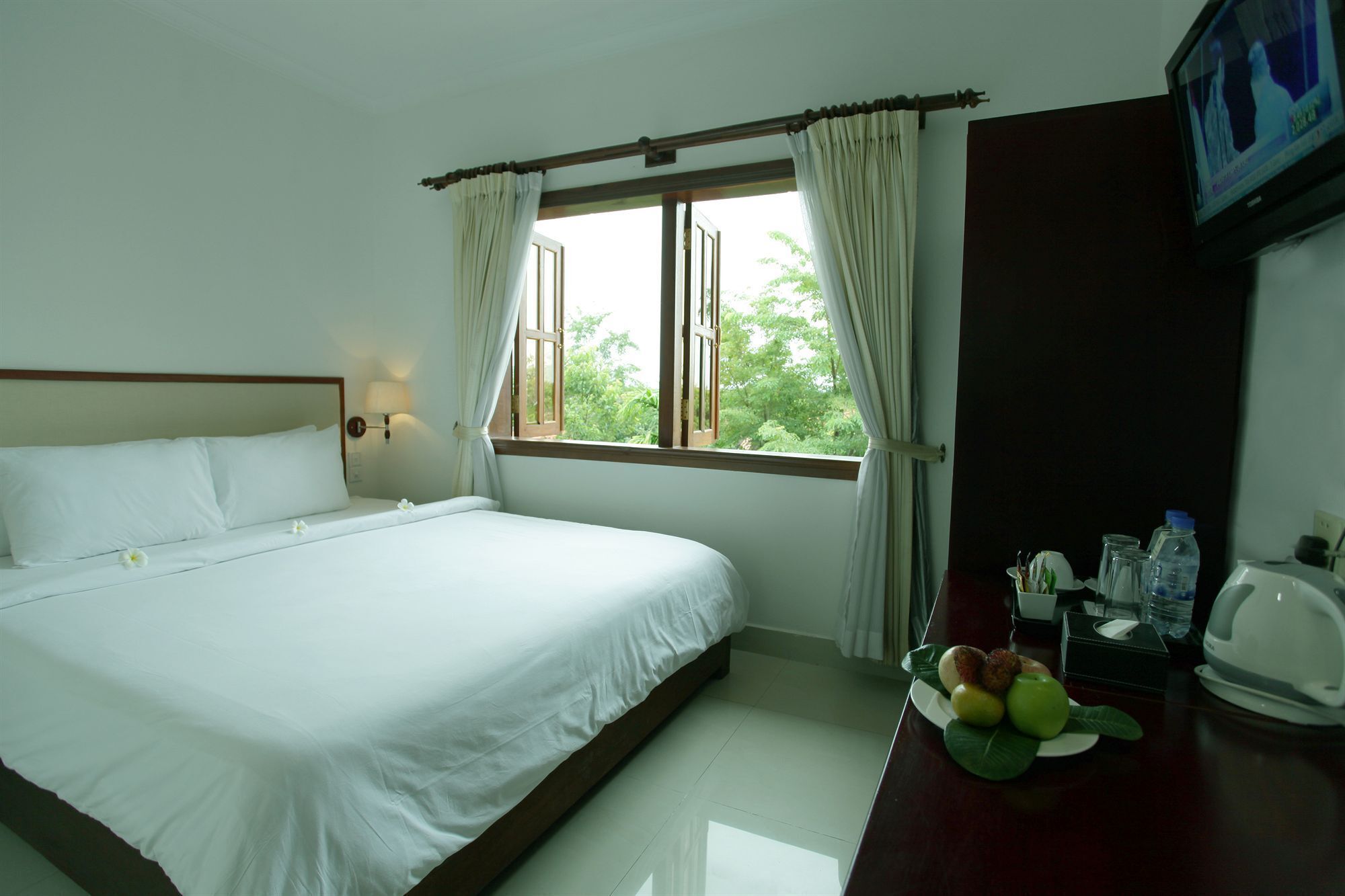 A hotel room with a large bed, a window with curtains, a desk with a plate of fruit, and a television on the wall.