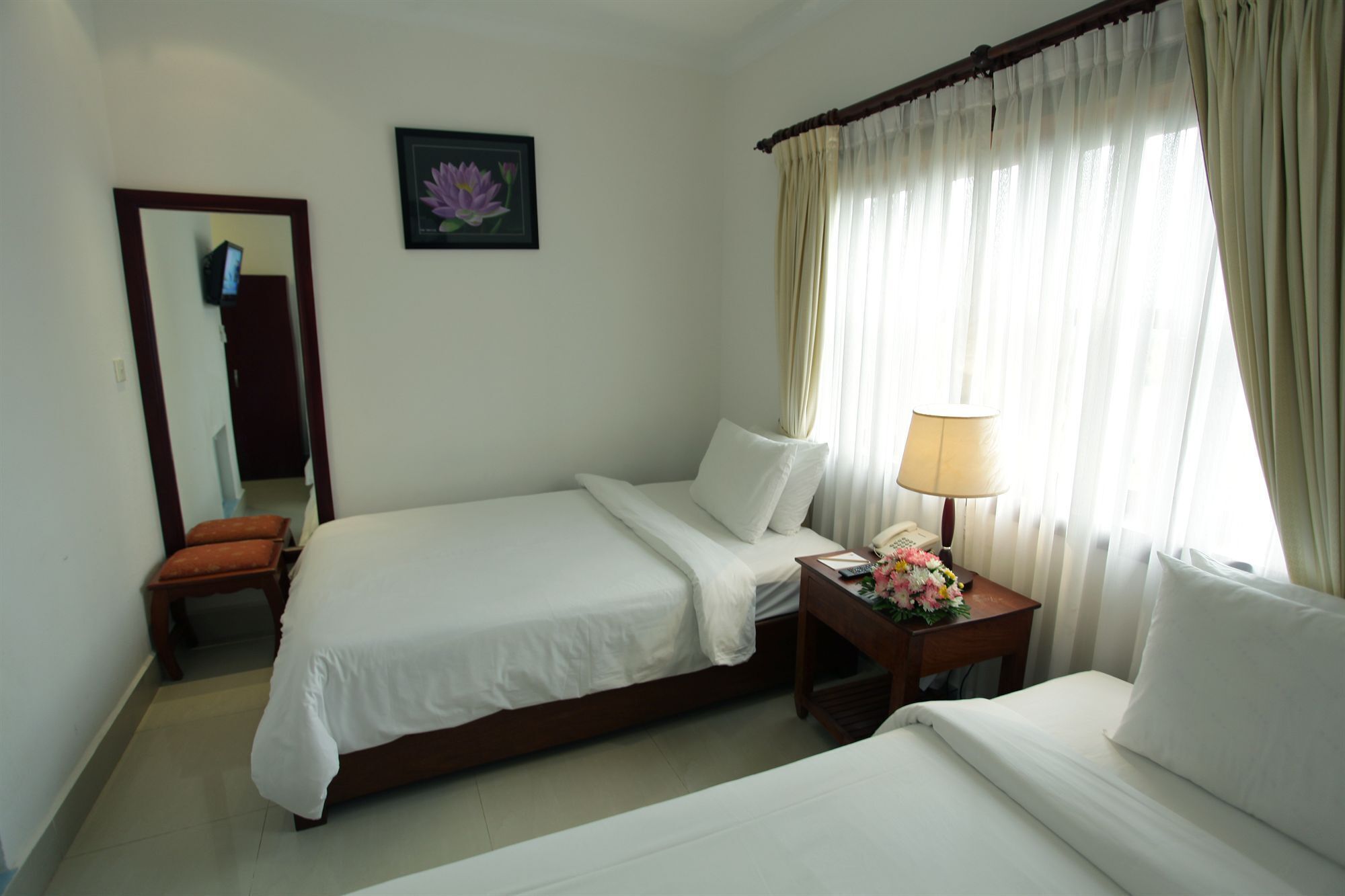 A hotel room with two single beds, a bedside table with a lamp and flowers, a mirror, and a framed picture on the wall.