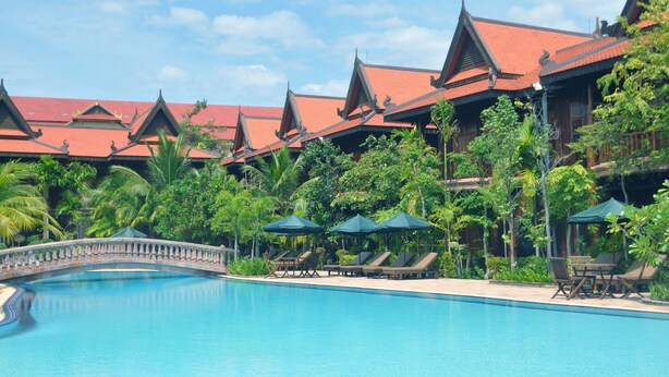 A swimming pool with lounge chairs and umbrellas, surrounded by lush greenery and traditional architecture. - Sokhalay Angkor Inn (Siem Reap)