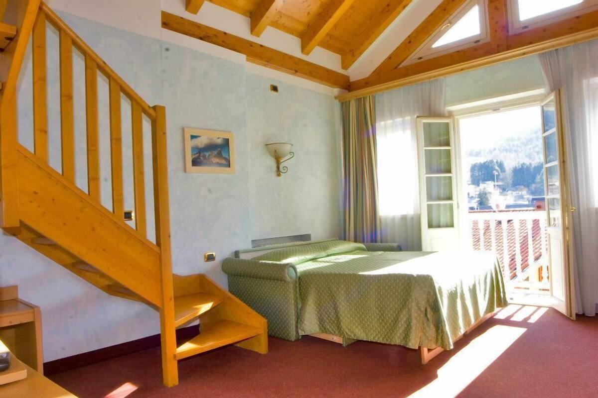 A bedroom with a wooden staircase, a bed with a green bedspread, a wall-mounted light fixture, and a window with a view of a mountain.