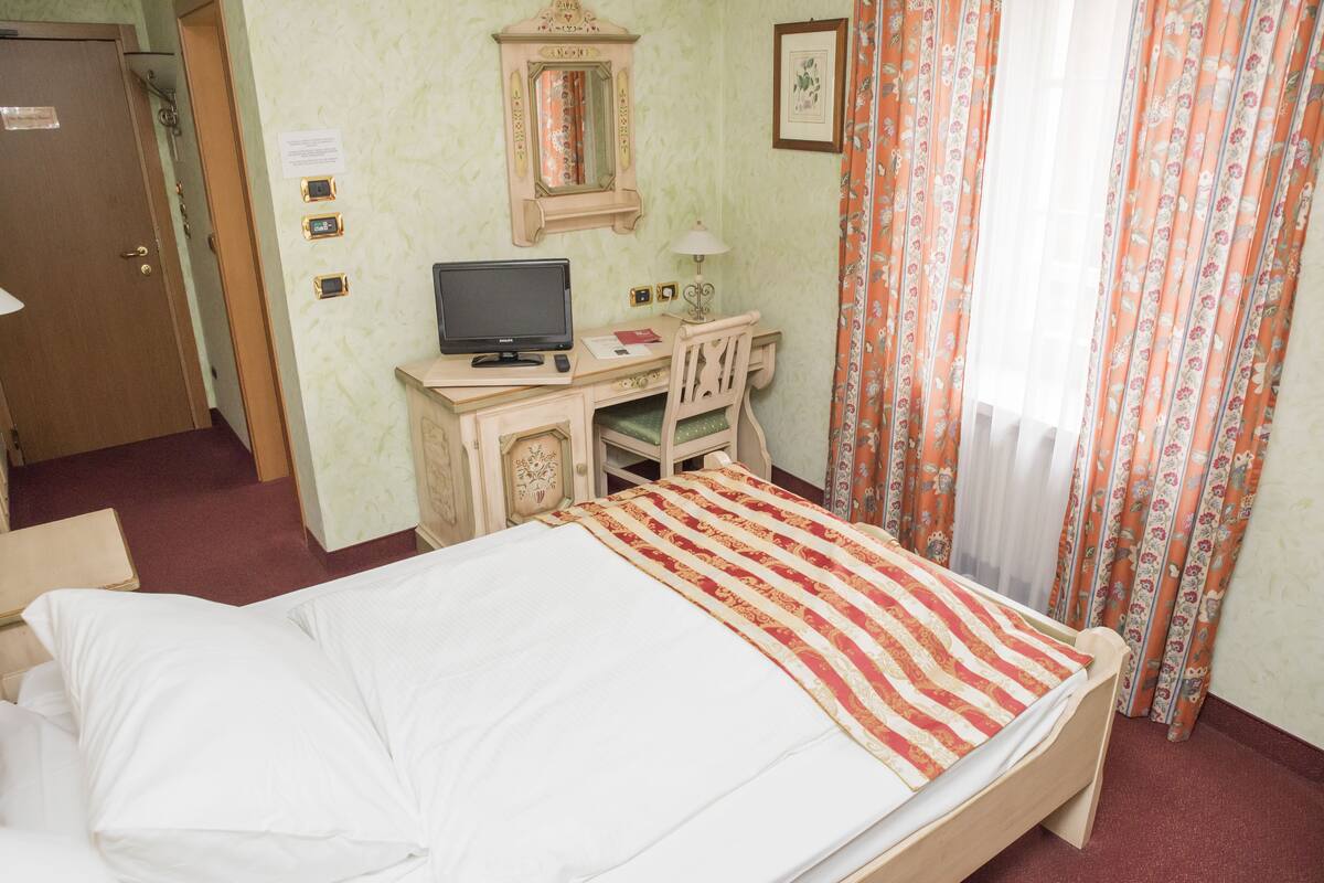 A hotel room with a bed, a desk with a computer, a mirror, and a window with curtains.