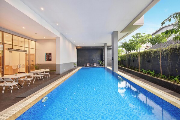 Una piscina interior moderna con agua cristalina azul, rodeada de sillones de descanso y un muro de cristal.