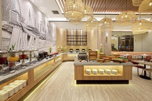 Un bar à petit-déjeuner moderne, agrémenté d’une fresque représentant un paysage urbain, de divers distributeurs de nourriture et de boissons, et d’un îlot central où sont disposées des assiettes.