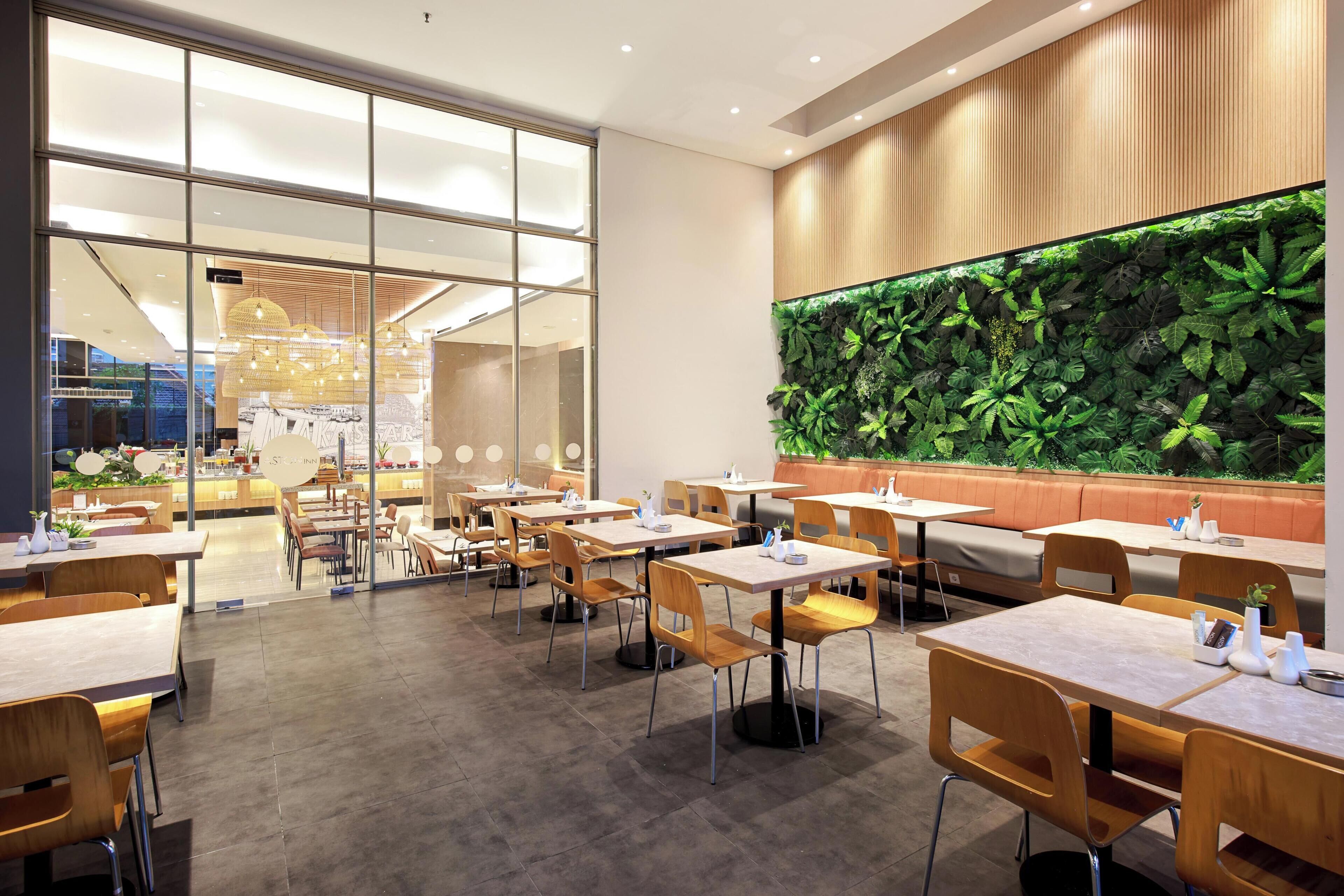 Un restaurante moderno con paredes de vidrio, mesas de madera y una pared verde con plantas.
