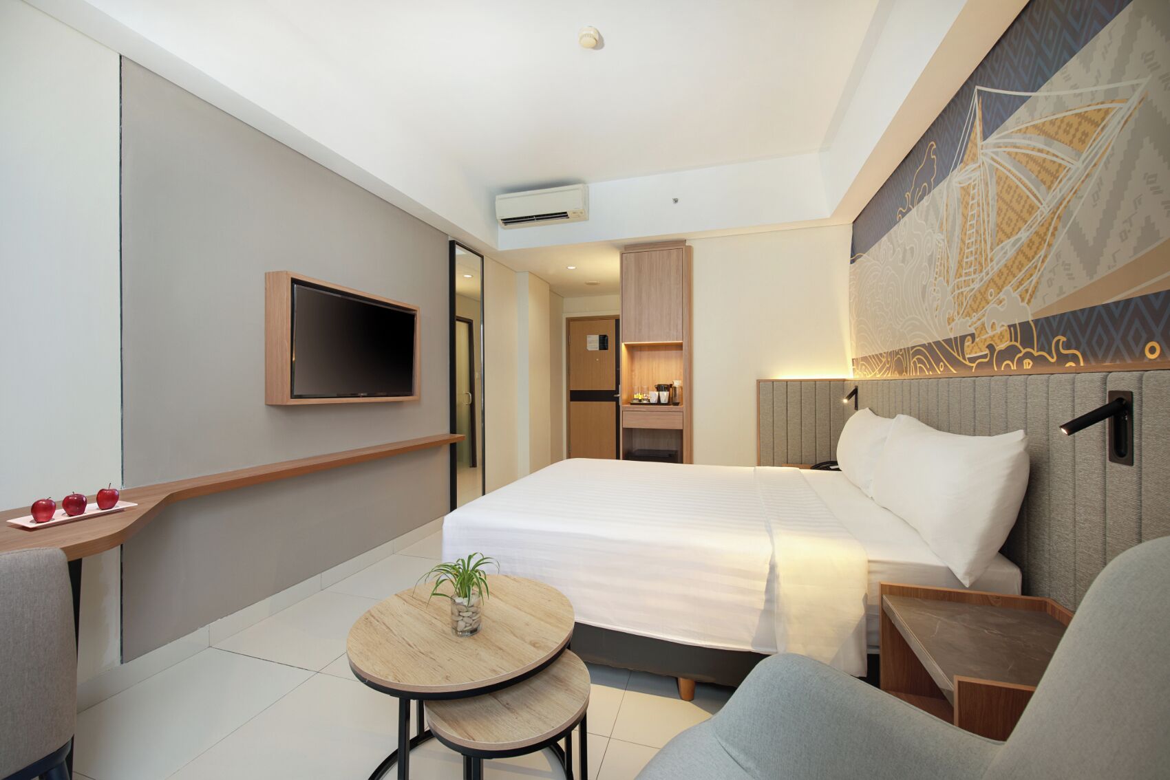 Een moderne hotelkamer met een groot bed, een aan de muur bevestigde televisie, een klein tafeltje met een plant en een zithoek met een fauteuil.