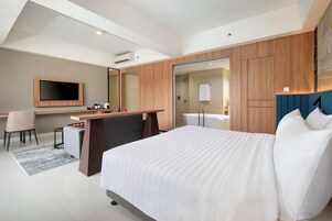 A modern hotel room with a large bed, a desk with a chair, a flat-screen TV, and a bathroom with a bathtub.