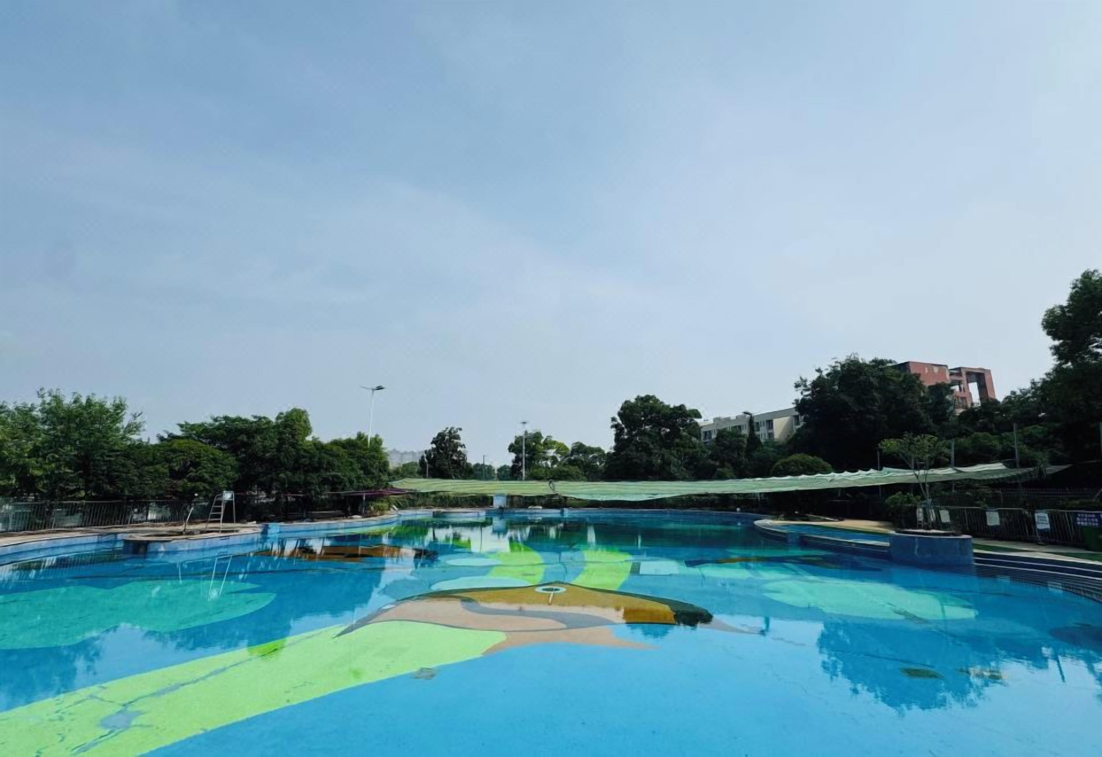 A large outdoor swimming pool with a clear blue water surface, surrounded by trees and a building in the background.