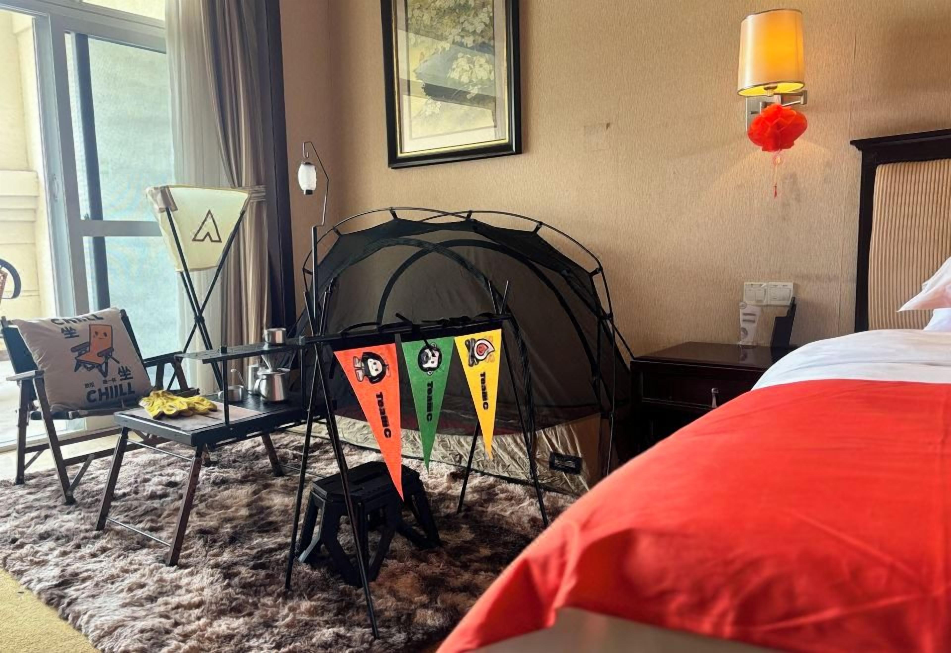 A hotel room with a bed, a chair, a small table, a tent, and a window with curtains.