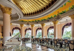 Um amplo hall com um piano de cauda, colunas decoradas, lustres e uma grande janela com vista para o verde.