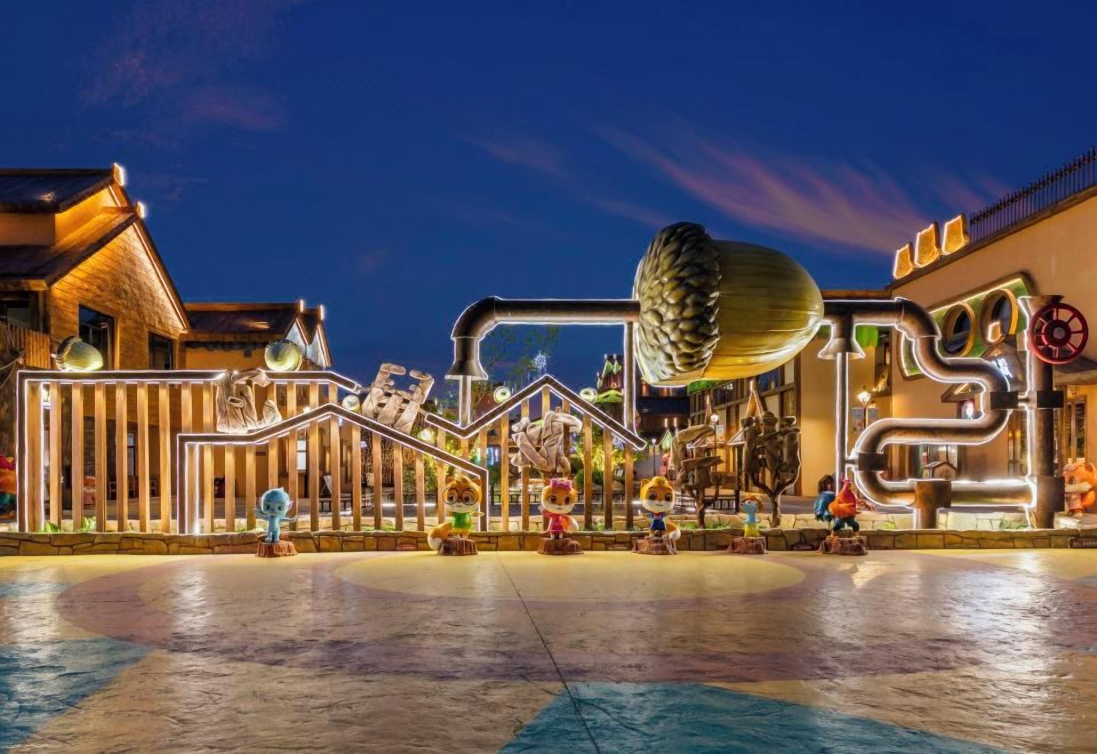 A nighttime view of a themed park entrance with illuminated structures and a large, central decorative element.