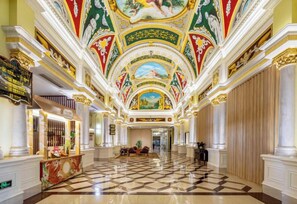 Un lobby de hotel imponente con piso de cuadros, columnas ornamentadas y un mural en el techo.