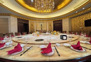 Una mesa redonda dispuesta para una cena formal, con una araña colgante, un papel tapiz decorativo y un televisor en la pared.