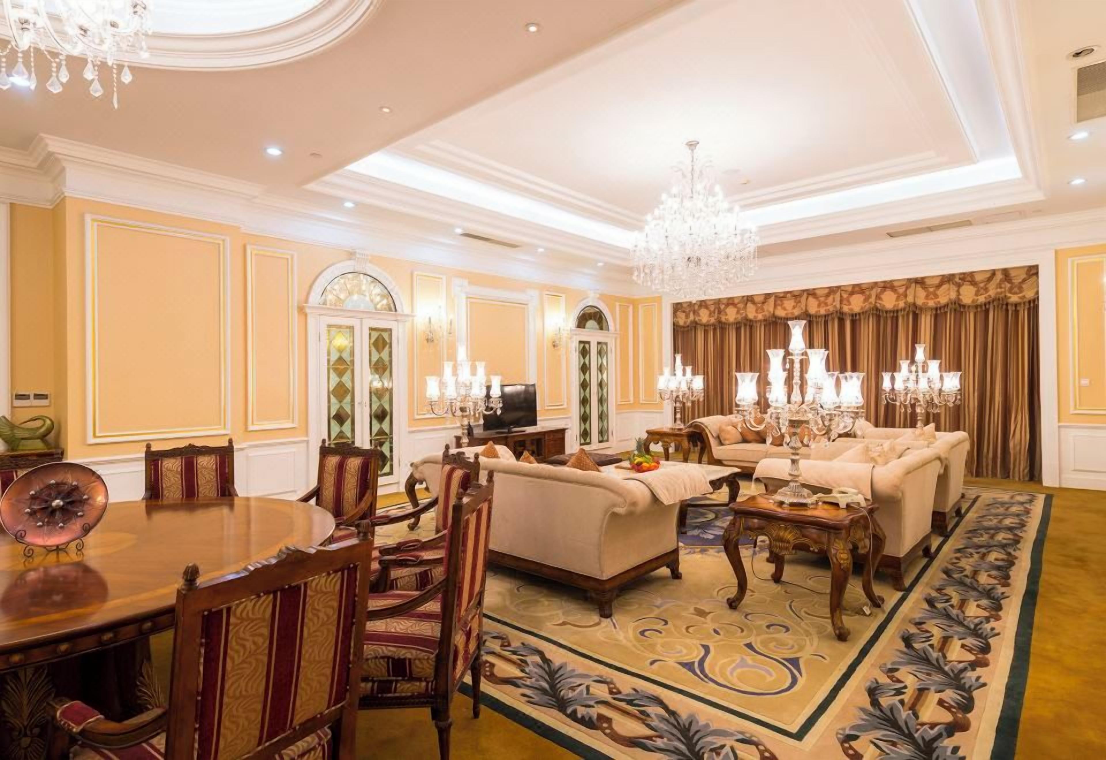 A spacious living room with a dining area, ornate chandeliers, and a patterned carpet.