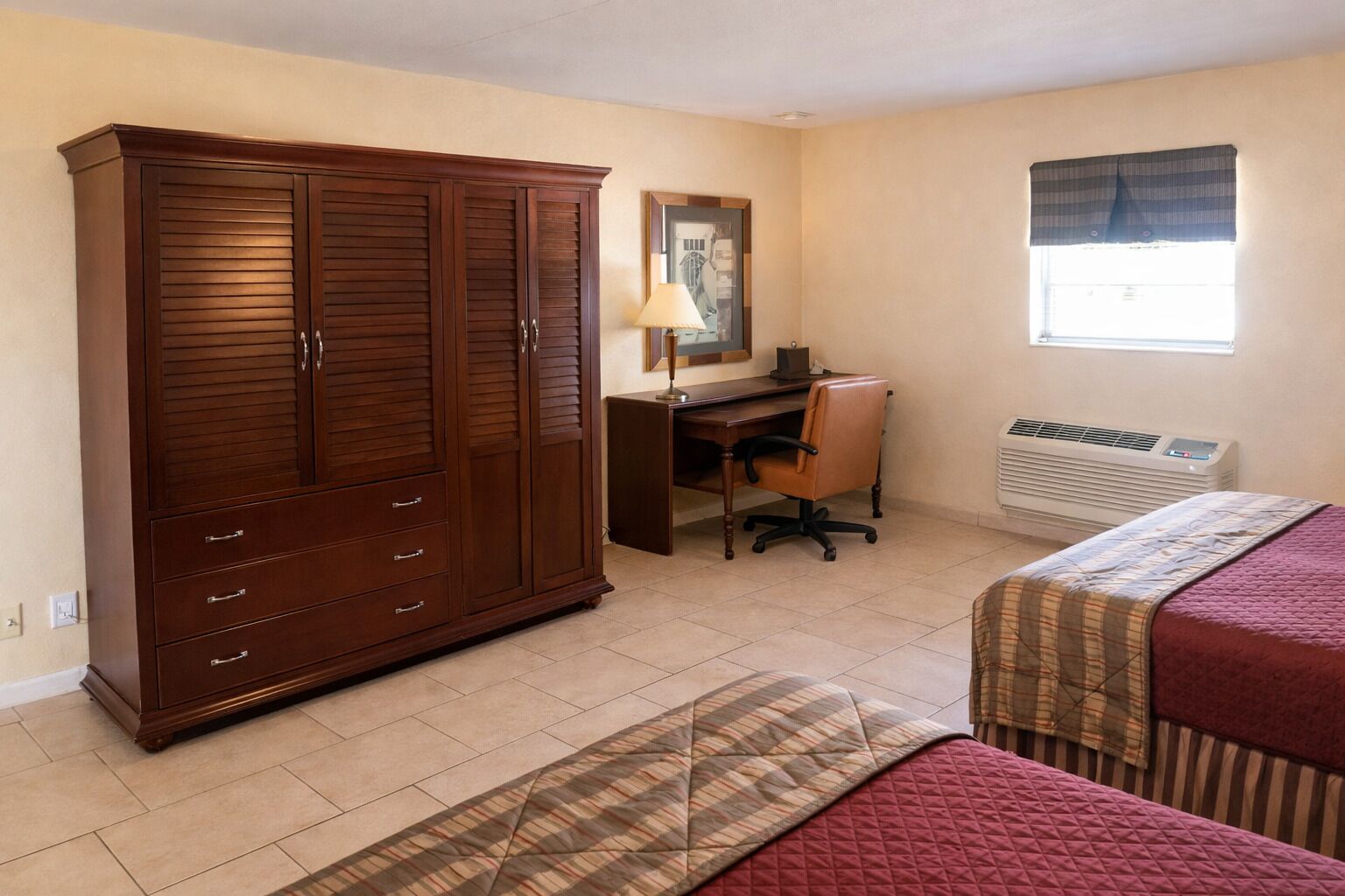 Une chambre avec une grande armoire en bois, un bureau avec une chaise, un lit avec une couette et une fenêtre avec un rideau.