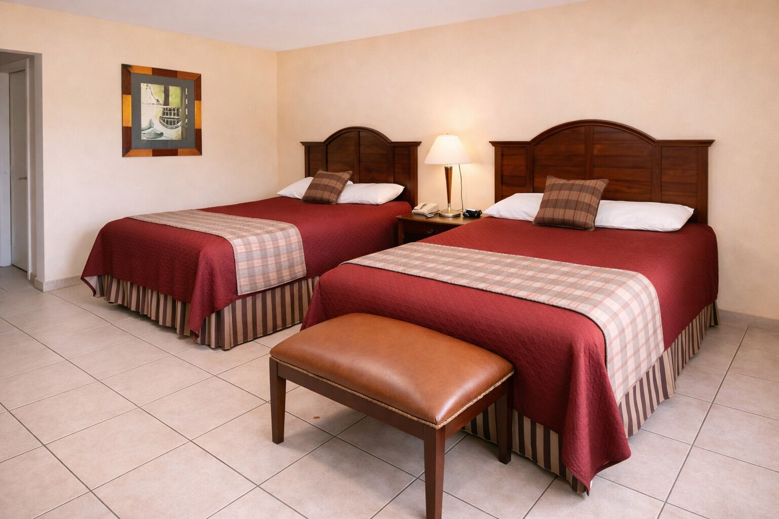 A hotel room with two beds, a bench, a bedside table with a lamp, and a framed picture on the wall.