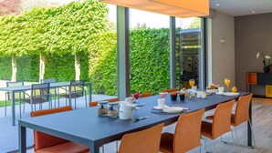 A modern dining area with a long table, orange chairs, and a glass partition. - B&B Antares (Bruges)
