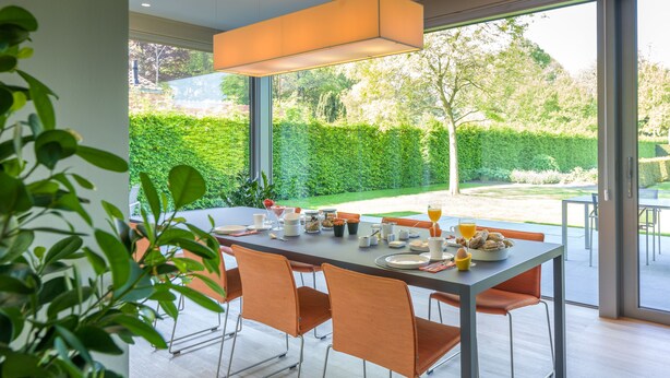 A modern dining area with a large table set for breakfast, large windows offering a view of a well-maintained garden. - B&B Antares (Bruges)