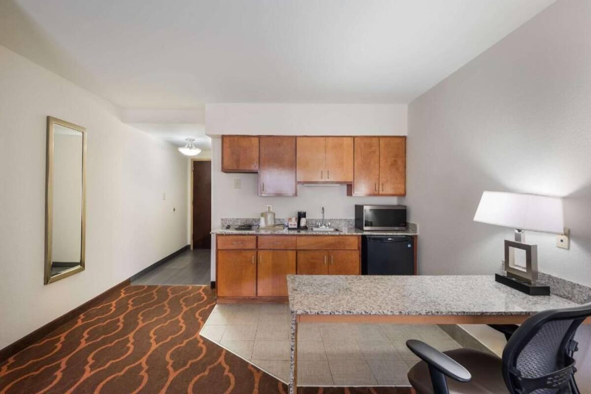 A compact hotel room with a kitchenette, a dining area with a table and chairs, a large mirror, and a patterned carpet.