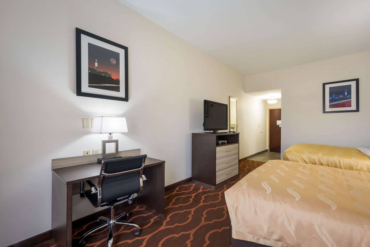 A hotel room with a desk, chair, television, bed, and two framed pictures on the walls.
