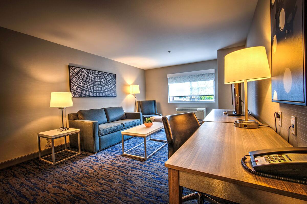 A hotel room with a wooden desk, a blue sofa, a small table with a lamp, and a painting on the wall.