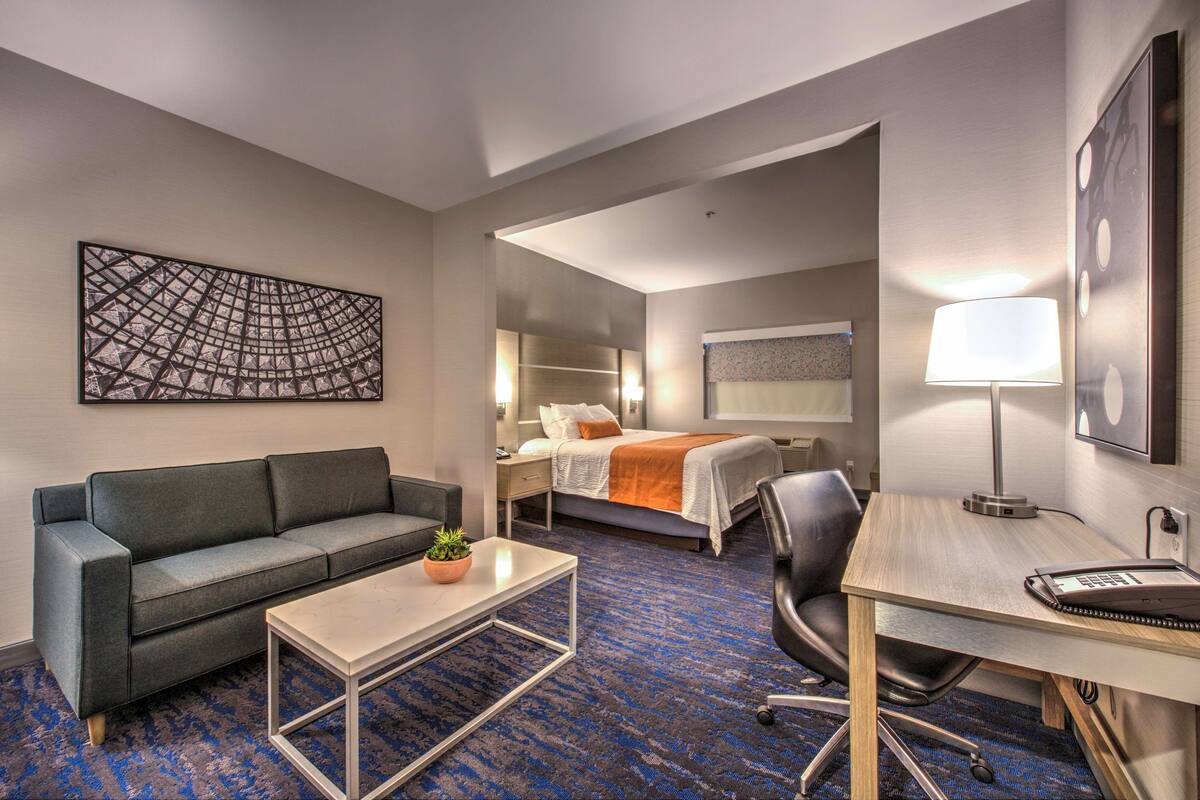 A hotel room with a sofa, a small table, a desk with a lamp, a bed with an orange blanket, and a large abstract painting on the wall.