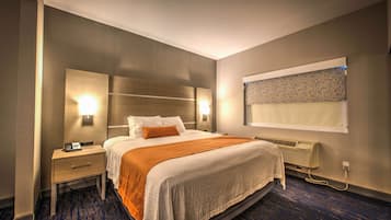 Une chambre d’hôtel avec un grand lit, une table de chevet, une lampe fixée au mur, un appareil de climatisation et une fenêtre avec un store à motifs.